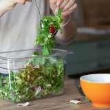 Detailreiches Produktfoto eines transparenten Gastronomiebehälters mit frischem Blattsalat, der mit Besteck in eine orangefarbene Schüssel gefüllt wird, Beispiel für professionelle Produktfotografie im Bereich Küchenzubehör und Gastronomiebedarf.